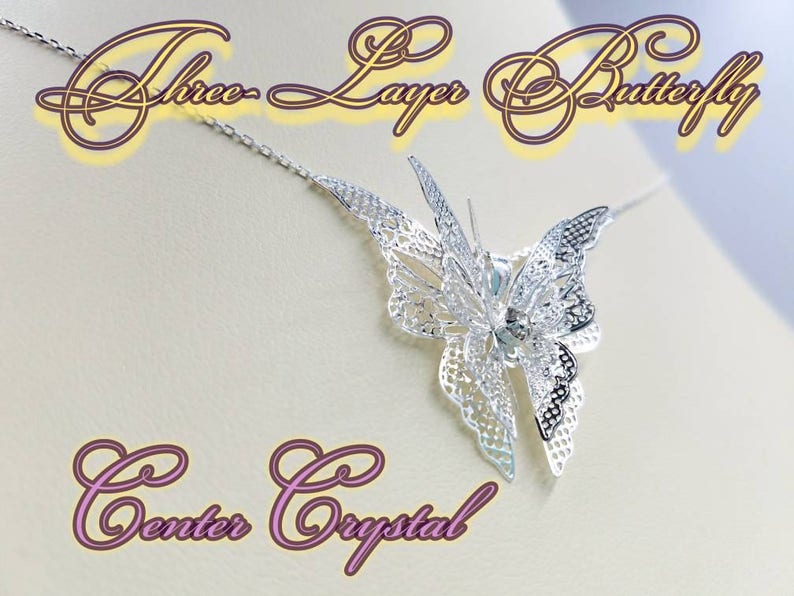 May include: Silver butterfly necklace with three layers of intricate detail and a clear crystal in the center. The necklace is on a white background.