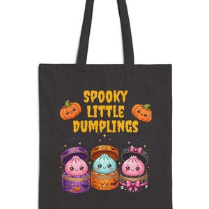 Puede incluir: Una bolsa de tela negra con un estampado gráfico que incluye el texto "SPOOKY LITTLE DUMPLINGS" en amarillo. El diseño muestra dos calabazas naranjas y tres dumplings coloridos y adorables en vaporeras de bambú con murciélagos y lazos.