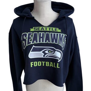 Puede incluir: Sudadera con capucha corta azul marino con el logotipo de los Seattle Seahawks en blanco y verde. La sudadera tiene una capucha con cord&oacute;n y un dise&ntilde;o de cuello en V. La palabra "SEATTLE" est&aacute; encima del nombre del equipo y "FOOTBALL" debajo del logotipo.