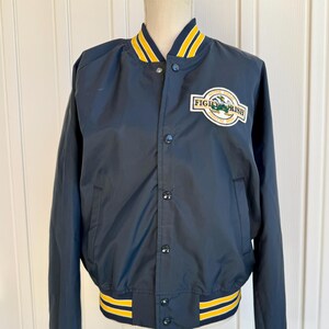 Puede incluir: Chaqueta bomber azul marino con detalles de rayas amarillas y blancas en el cuello, los puños y el dobladillo. Cuenta con un parche con el texto "FIGHTING IRISH" y un logotipo de duende verde. Cierre de botones y dos bolsillos delanteros.