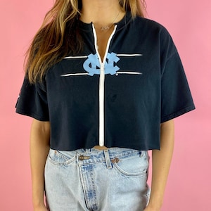 Op de afbeelding: Een zwart gecropped t-shirt met een witte rits aan de voorkant. Het shirt heeft een lichtblauw UNC-logo met witte strepen aan weerszijden van het logo.