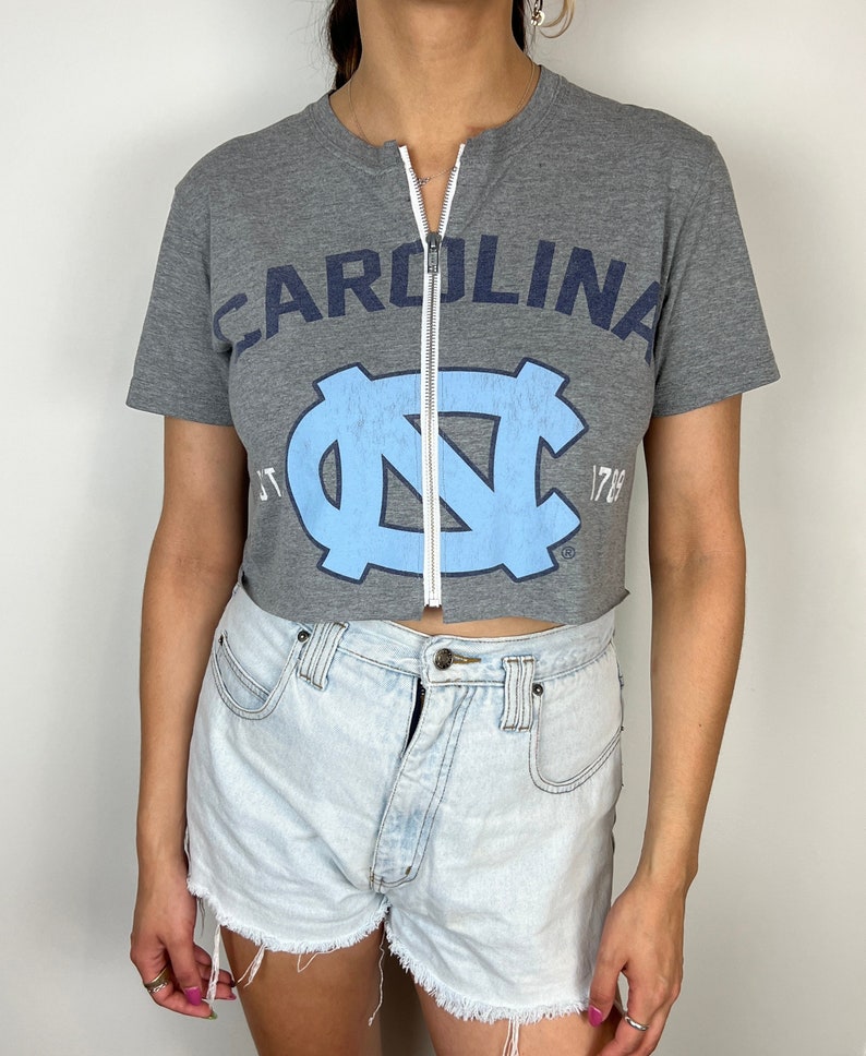 Puede incluir: Una camiseta gris recortada con un logotipo UNC azul y blanco y el texto "CAROLINA" encima. La camiseta tiene una cremallera en la parte delantera.