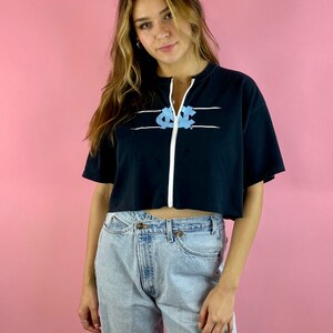 Op de afbeelding: Een zwart gecropped zip-up sweatshirt met een lichtblauwe afbeelding van een college logo. Het sweatshirt heeft witte strepen over de borst. Het model draagt ​​lichtblauwe jeans.