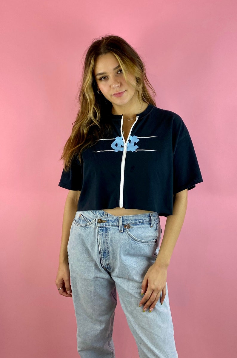 Op de afbeelding: Een zwart gecropped zip-up sweatshirt met een lichtblauwe afbeelding van een college logo. Het sweatshirt heeft witte strepen over de borst. Het model draagt ​​lichtblauwe jeans.