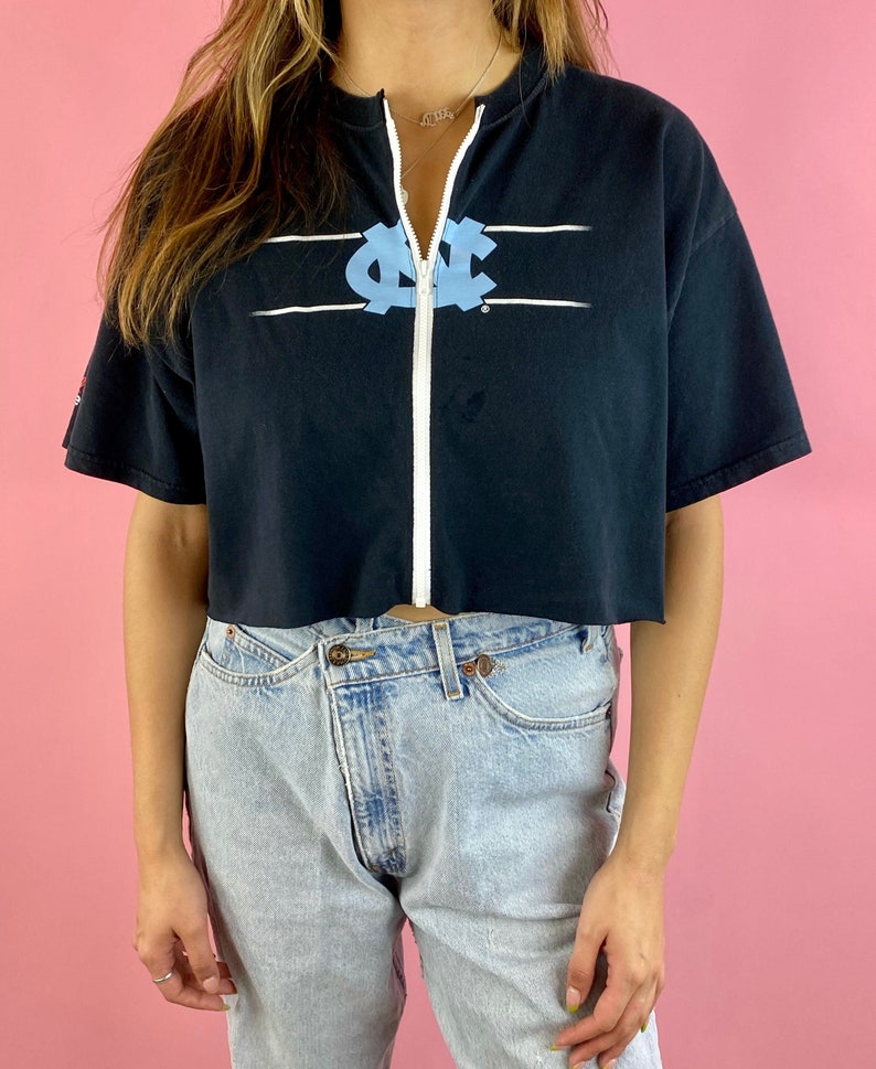 Op de afbeelding: Een zwart gecropped t-shirt met een witte rits aan de voorkant. Het shirt heeft een lichtblauw UNC-logo met witte strepen aan weerszijden van het logo.