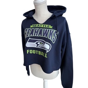 Puede incluir: Sudadera corta azul marino con el logo de los Seattle Seahawks. La sudadera con capucha presenta el nombre y el logotipo del equipo en blanco y verde ne&oacute;n. La palabra "FOOTBALL" est&aacute; impresa debajo del logotipo. La sudadera tiene un cord&oacute;n y un cuello en V.