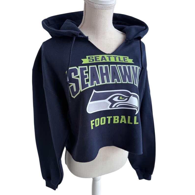 Puede incluir: Una sudadera corta azul marino con el logotipo y el texto de los Seattle Seahawks. El dise&ntilde;o incluye el nombre del equipo y una cabeza de &aacute;guila estilizada en blanco, verde y azul. La palabra "FOOTBALL" est&aacute; impresa debajo.