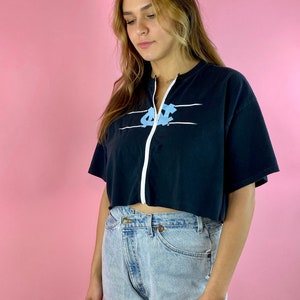 Op de afbeelding: Een zwart gecropped t-shirt met een witte rits en een blauwe en witte afbeelding van de letters "UNC" op de voorkant. Het shirt wordt gedragen door een persoon met lang blond haar en wordt gecombineerd met lichtblauwe jeans.