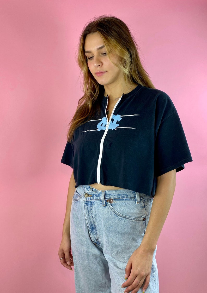 Op de afbeelding: Een zwart gecropped t-shirt met een witte rits en een blauwe en witte afbeelding van de letters "UNC" op de voorkant. Het shirt wordt gedragen door een persoon met lang blond haar en wordt gecombineerd met lichtblauwe jeans.