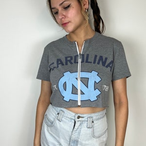 Puede incluir: Una camiseta gris recortada con un logotipo azul y blanco de la Universidad de Carolina del Norte y la palabra "CAROLINA" impresa en la parte delantera. La camiseta tiene una cremallera en la parte delantera.