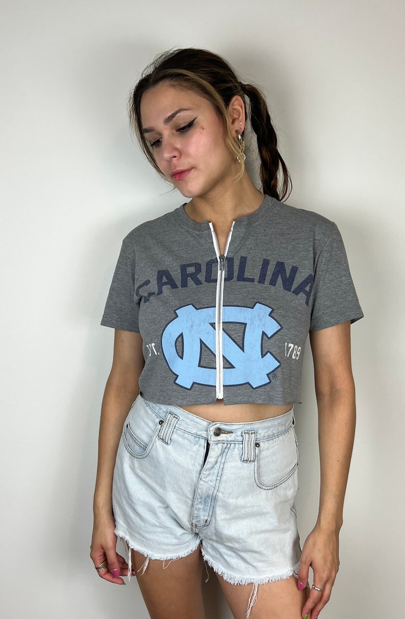 Puede incluir: Una camiseta gris recortada con un logotipo azul y blanco de la Universidad de Carolina del Norte y la palabra "CAROLINA" impresa en la parte delantera. La camiseta tiene una cremallera en la parte delantera.