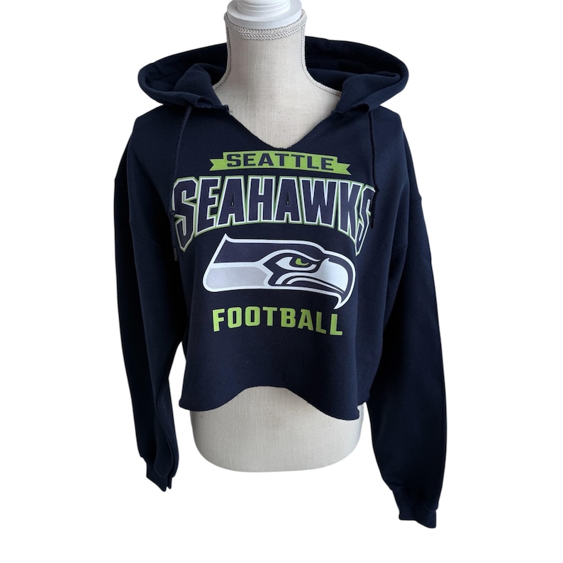 Puede incluir: Sudadera con capucha corta azul marino con el logotipo de los Seattle Seahawks en blanco y verde. La sudadera tiene una capucha con cord&oacute;n y un dise&ntilde;o de cuello en V. La palabra "SEATTLE" est&aacute; encima del nombre del equipo y "FOOTBALL" debajo del logotipo.