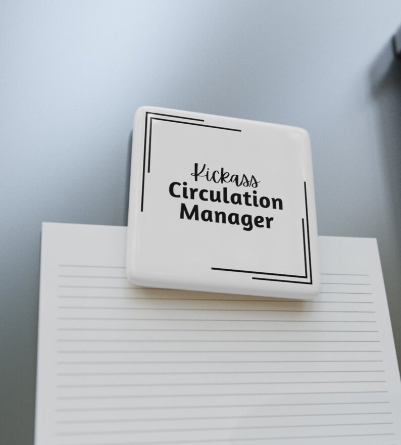 Square white porcelain magnet on a refrigerator holding paper. Text on the white magnet reads Kickass [italics] Circulation Manager [non-italics] with double corner brackets in the upper left and lower right corner.