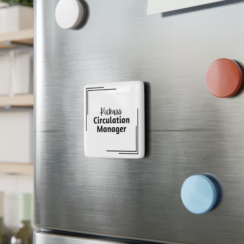 Square white porcelain magnet on a refrigerator surrounded by round colored magnets. Text on the white magnet reads Kickass [italics] Circulation Manager [non-italics] with double corner brackets in the upper left and lower right corners.