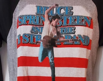 May include: A grey and black baseball style t-shirt with a vintage image of Bruce Springsteen and the E Street Band. The image features Springsteen playing a guitar in front of an American flag.