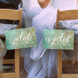 May include: Two wooden chairs with a vintage map design sign that says "pilot" and "co-pilot" hanging from the back of each chair. The chairs are set up for a wedding reception.