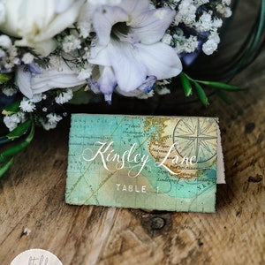 May include: A vintage-style map place card with a compass rose and the text "Kinsley Lane Table 1". The place card is on a wooden surface with a bouquet of white and purple flowers.