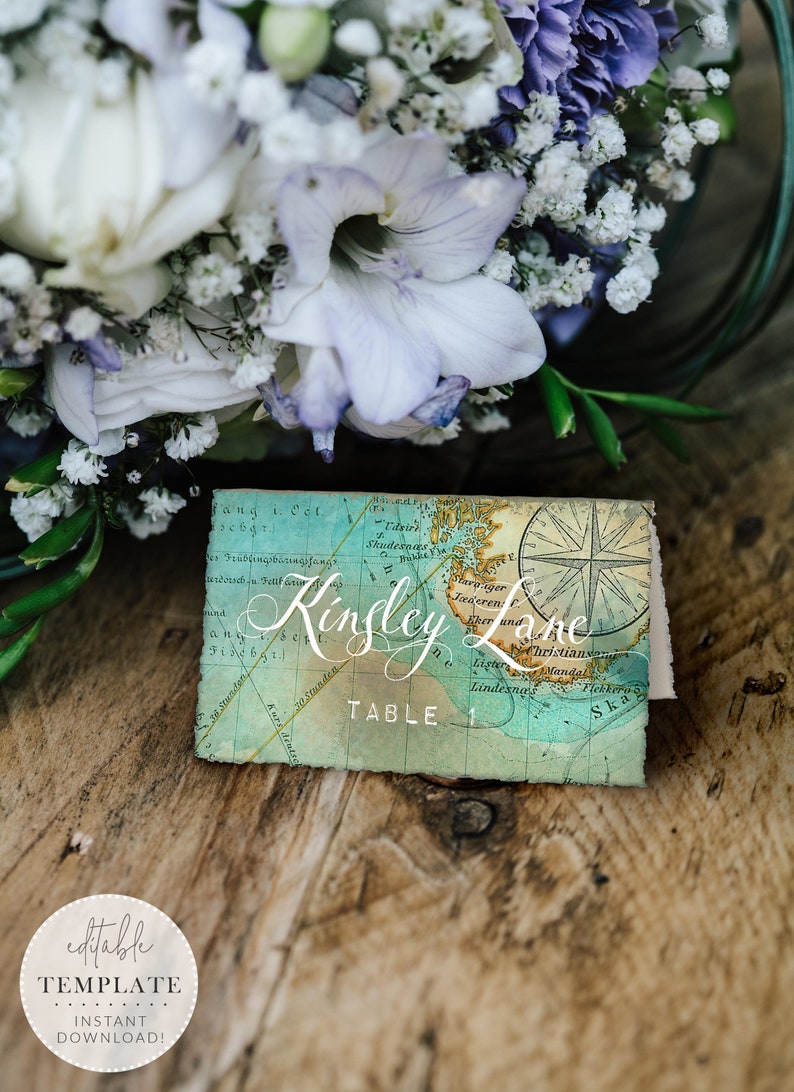 May include: A vintage-style map place card with a compass rose and the text "Kinsley Lane Table 1". The place card is on a wooden surface with a bouquet of white and purple flowers.