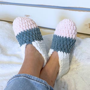 May include: Cozy, handmade slippers in a soft, textured knit. The slippers feature a color block design with sections of pink, gray, and white. The open-toe design and plush material suggest comfort and relaxation. Perfect for lounging at home.