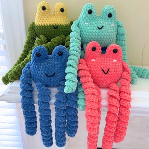 May include: Four handmade crochet frog plushies in various colors: green and yellow, teal, blue, and pink. Each frog has large black eyes and a smiling mouth. The frogs have long, curly legs.