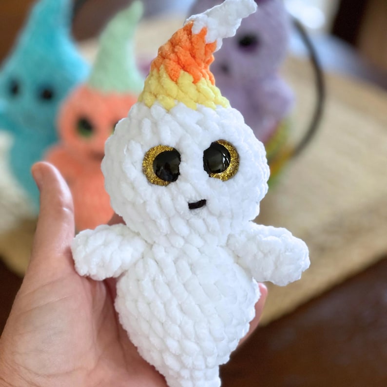 May include: White crocheted ghost with a candy corn hat and sparkly gold eyes. The ghost is holding its arms out and has a small smile.