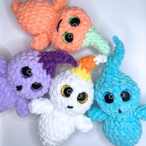 May include: Four crocheted ghost plushies in different colors. The ghosts are white, purple, orange, and blue. Each ghost has googly eyes and a stitched-on smile.