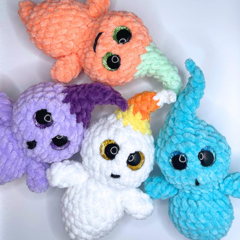 May include: Four crocheted ghost plushies in different colors. The ghosts are white, purple, orange, and blue. Each ghost has googly eyes and a stitched-on smile.