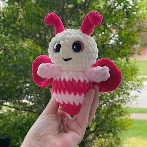 May include: A crocheted pink and white butterfly plush toy with sparkly eyes. The toy is held in a hand against a green background.
