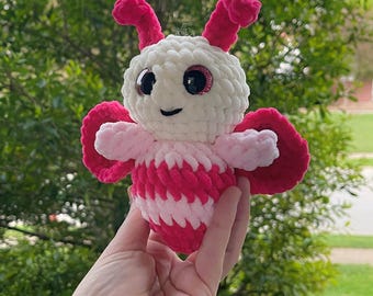 Peut inclure: Un jouet en peluche en forme de papillon crocheté en rose et blanc avec des yeux scintillants. Le jouet est tenu dans une main sur un fond vert.