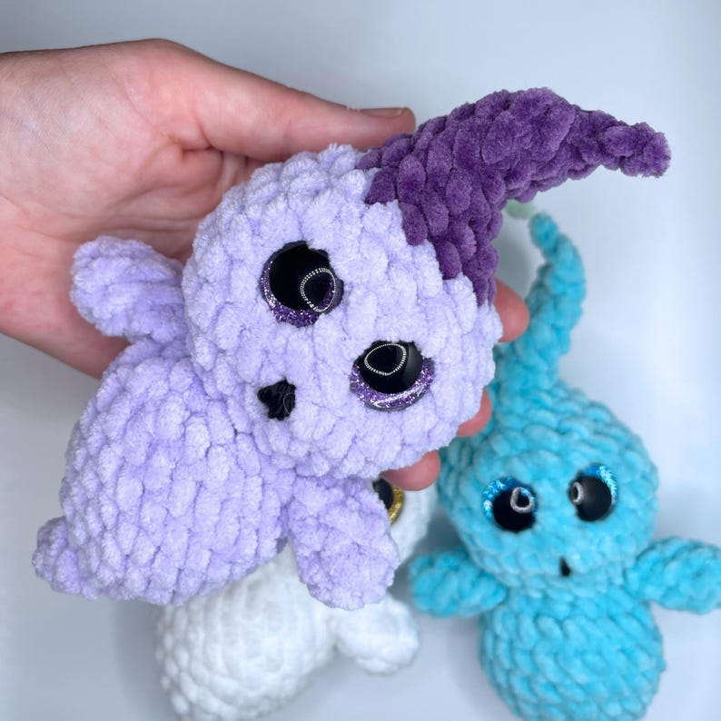 May include: Three crocheted plush toys in the shape of ghosts. The first ghost is purple with a purple horn, the second ghost is white, and the third ghost is blue.