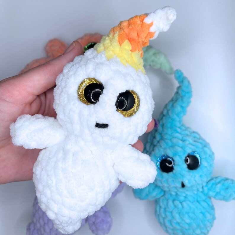May include: Three crocheted plush toys. One is a white ghost with a rainbow yarn hat, another is a light blue ghost, and the third is a purple ghost.