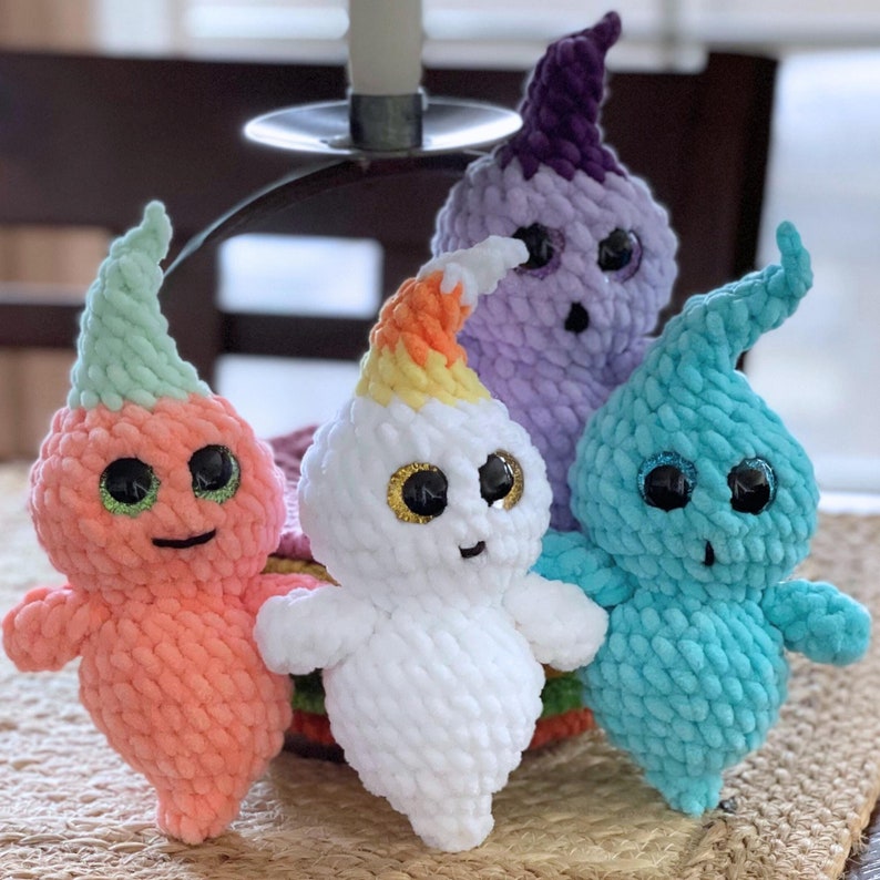 May include: Four crocheted ghost plushies in different colors. The ghosts are made of yarn and have googly eyes. The ghosts are sitting on a table.