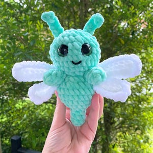 Puede incluir: Un juguete de peluche verde y blanco tejido a crochet con forma de mariposa con grandes ojos negros. El juguete tiene una boca sonriente y alas blancas.