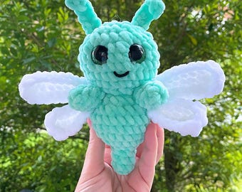May include: A crocheted green and white plush toy shaped like a butterfly with large black eyes. The toy has a smiling mouth and white wings.
