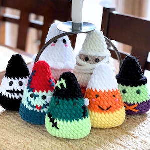 Puede incluir: Ocho adornos de maíz dulce de crochet en varios colores, incluyendo negro, blanco, verde, rojo, naranja y amarillo. Los adornos están dispuestos en un círculo sobre una superficie tejida.