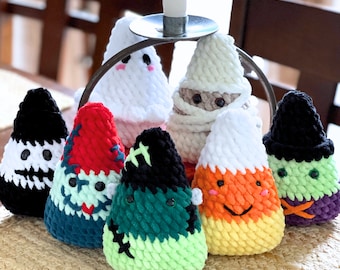 May include: Eight crocheted candy corn ornaments in various colours, including black, white, green, red, orange, and yellow. The ornaments are arranged in a circle on a woven surface.