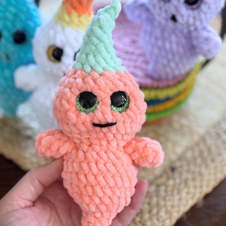 May include: A crocheted plush toy shaped like a ghost with a green and white crocheted hat. The ghost has large, glittery green eyes and a black smile. The toy is peach colored.