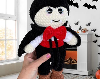 May include: A handmade plush vampire doll with a black cape, red bow tie, and brown boots. The doll has a white face with rosy cheeks, large eyes, and a small smile. The doll is a Halloween-themed item.