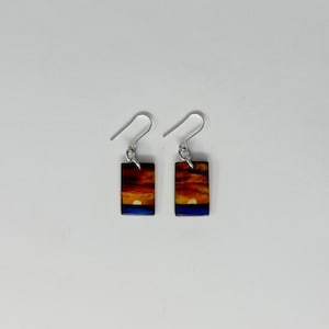 May include: A pair of silver dangle earrings with a rectangular sunset scene. The scene features a yellow sun setting over a blue ocean with orange and red clouds.