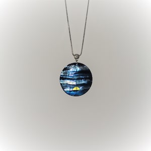 May include: A round pendant necklace with a silver chain. The pendant is a dark blue and black iridescent circle with a yellow and gold design. The necklace is displayed against a white background.