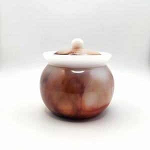 May include: A small, round jar with a lid. The jar is a mix of brown and white colors, with a white rim and a small, round knob on top of the lid. The jar has a glossy finish and appears to be made of a smooth material.