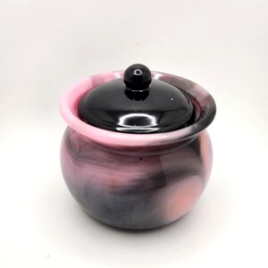 May include: A small, round jar with a lid. The jar has a swirling pattern of pink, black, and white. The lid is black and has a small, round knob on top. The jar is sitting on a white surface.