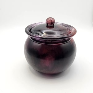 May include: A small, round, purple glass jar with a matching lid and a small knob on top. The jar has a wide opening and a slightly flared rim. The glass has a marbled appearance, with darker and lighter shades of purple.