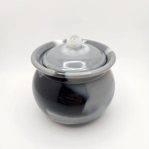 May include: A small, round, lidded jar with a glossy finish. The jar is primarily black and gray, with a white knob on the lid. The jar has a smooth, rounded shape and appears to be made of ceramic or glass.