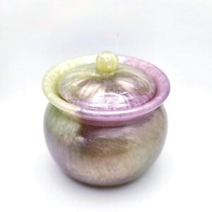 Può includere: Un piccolo barattolo rotondo con coperchio. Il barattolo è un mix di colori, tra cui viola, verde e argento. Il coperchio ha un piccolo pomello in cima. Il barattolo è probabilmente fatto di resina o un materiale simile.