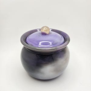 Puede incluir: Un pequeño tarro redondo con tapa. El tarro tiene un degradado de color, que va del morado oscuro al plateado. La tapa es de un tono morado más claro con un pequeño pomo redondo y claro en la parte superior. El tarro probablemente se usa para guardar cosas.