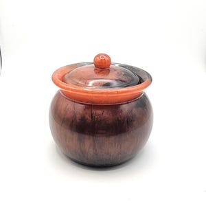 Può includere: Un piccolo vaso rotondo con coperchio dall'aspetto di legno marrone rossiccio. Il coperchio ha un bordo arancione rossiccio e un piccolo pomello rotondo. Il corpo del vaso è di una tonalità di marrone più scura, con finitura liscia e lucida.