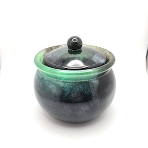 Peut inclure: Petit pot rond en céramique avec couvercle. Le pot est émaillé dans des tons de vert et de bleu, avec une finition brillante. Le couvercle a un petit bouton rond. Le pot est probablement utilisé pour ranger de petits objets ou comme pièce décorative.