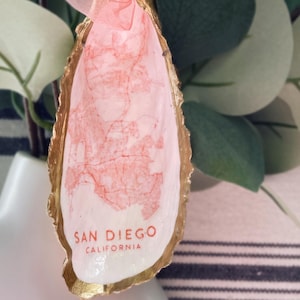 Könnte beinhalten: Ein rosa-goldener Austernmuschel-Anhänger mit dem Aufdruck "SAN DIEGO CALIFORNIA" auf der Innenseite.