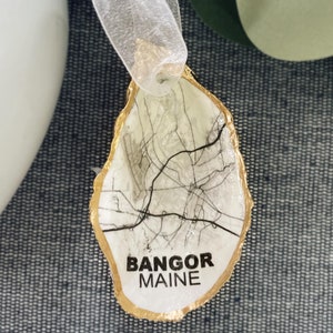 May include: Gold-painted oyster shell ornament with a black and white map of Bangor, Maine printed on the inside. The ornament has a white ribbon attached for hanging.